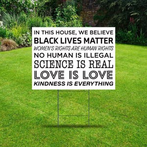 May include: A black and white yard sign with the text "In this house, we believe Black Lives Matter, Women's Rights are Human Rights, No Human is Illegal, Science is Real, Love is Love, Kindness is Everything."