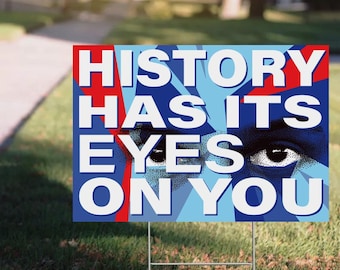 May include: A blue and red yard sign with the words "HISTORY HAS ITS EYES ON YOU" in bold white letters. The sign features an illustration of an eye. The sign is supported by a metal stake.