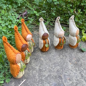 Puede incluir: Una fila de siete figuras de muñecas de fieltro con sombreros puntiagudos. Las muñecas están dispuestas en línea, con los sombreros alternando entre naranja y beige. Cada muñeca tiene una cabeza de madera y un collar de cuentas con abalorios de hojas verdes.