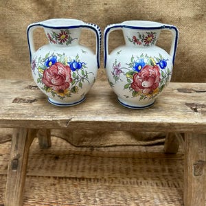 May include: Two small white ceramic vases with blue and red floral designs and blue trim. The vases have two handles each and are sitting on a wooden surface.