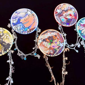 Puede incluir: Una colección de cinco pines redondos de personajes de estilo anime con detalles de cadena plateada. Cada pin presenta una ilustración de personaje diferente con colores vibrantes y detalles en forma de estrella. Las cadenas están adornadas con estrellas, lunas y pequeñas cuentas azules.