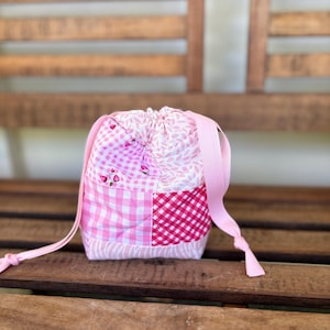 May include: A pink and red patchwork drawstring bag with a white background. The bag is made of fabric and has a pink ribbon drawstring.
