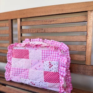 May include: A pink and white patchwork cushion with a ruffled edge. The cushion is made of different fabrics, including gingham and floral prints.