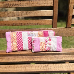 May include: Two pink and white patchwork pencil cases with floral and gingham patterns. The larger case has a rectangular shape, while the smaller one is also rectangular. The cases are resting on a wooden bench.