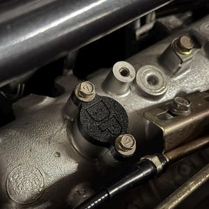 Puede incluir: Una pieza automotriz negra y texturizada con un logotipo, asegurada por tres pernos. La pieza está montada en un componente del motor plateado, con tubos metálicos y otros elementos en el fondo.