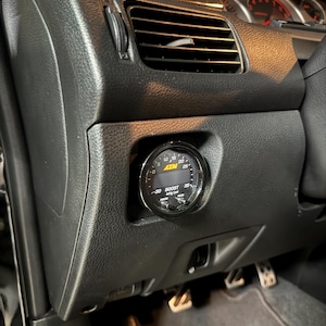 May include: A black dashboard with an AEM boost gauge installed. The gauge has a black face with white markings and a needle. The dashboard also features air vents and other controls.