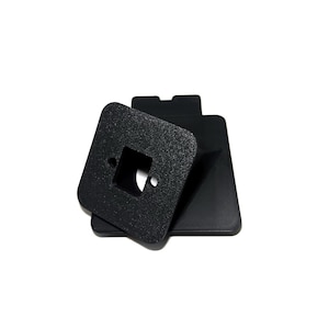 Puede incluir: Objeto negro, texturizado, de forma cuadrada con un recorte cuadrado central y un pequeño agujero circular. Descansa sobre una base negra rectangular más grande. Los objetos parecen ser de material mate.