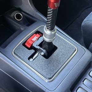 May include: A car gear shifter assembly featuring a carbon fiber shaft with a red top, a silver collar, and a black base. The mechanism includes a red metal component with the white letters IVK visible, set within a textured gray console plate.