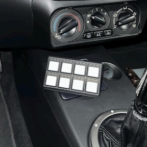 Puede incluir: Un panel de control rectangular negro con ocho botones cuadrados blancos dispuestos en dos filas de cuatro. El dispositivo está colocado en la consola central de un vehículo, situado directamente debajo de los diales e interruptores del climatizador.