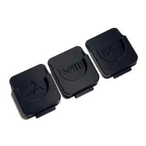 May include: Three black square-shaped plastic covers with rounded corners and a small tab at the bottom. Each cover features a distinct embossed symbol on the surface: a geometric diamond-like shape, the Roman numeral VIII, and the Roman numeral IX.