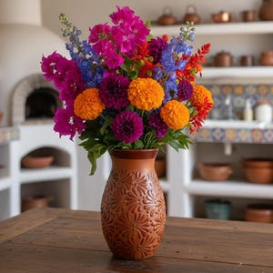May include: A colourful bouquet of flowers in a brown, textured vase. The arrangement includes orange, purple, blue, and pink blooms. The vase sits on a wooden table, with a brick arch and kitchen in the background.