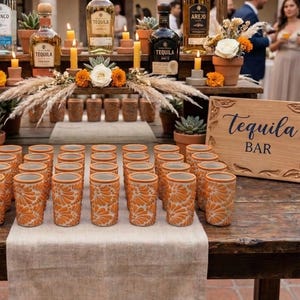Puede incluir: Un bar de tequila con botellas, vasos de chupito y un letrero de madera que dice "Tequila Bar". El bar está decorado con flores, velas y suculentas. Los vasos de chupito tienen un diseño floral naranja.