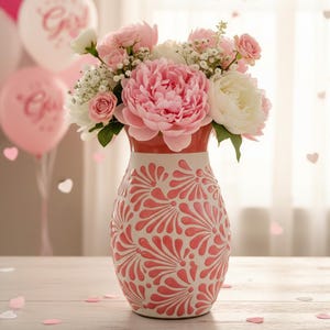 Puede incluir: Un arreglo floral con peonías rosas y flores blancas en un jarrón decorativo. El jarrón tiene un diseño floral rosa sobre fondo blanco. Globos rosas y una pancarta que dice "IT'S A GIRL" se ven en el fondo.