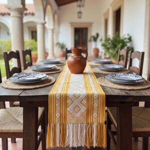 Puede incluir: Una mesa de comedor rústica al aire libre está preparada. La mesa está puesta con platos, manteles individuales tejidos y un camino de mesa con estampado amarillo y blanco. Una jarra marrón está en el centro. Sillas de madera rodean la mesa.