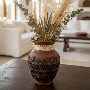 Op de afbeelding: Een bruine keramische vaas met ingewikkelde gesneden patronen en een geweven touwdetail rond de hals. De vaas bevat gedroogde planten en pampasgras, wat een natuurlijke, bohemien esthetiek creëert.