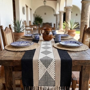 Puede incluir: Una mesa de comedor rústica para cuatro, con manteles individuales tejidos, platos y tazas con diseños azules y blancos, y una jarra marrón. Un camino de mesa geométrico negro, beige y blanco adorna la mesa de madera, creando un ambiente cálido.