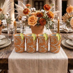 May include: A beautifully set table with floral centerpieces in terracotta pots, candles, and four orange patterned glasses with lime wedges. The table is set with plates and napkins, creating an inviting outdoor dining scene.