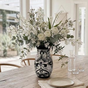 Pode incluir: Um vaso de cerâmica com padrão preto e branco preenchido com um grande arranjo de flores brancas e folhas de eucalipto verdes, colocado sobre uma mesa de jantar de madeira rústica ao lado de um prato pequeno, um guardanapo dobrado e uma jarra de vidro.