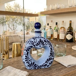 Op de afbeelding: Een lichtblauwe keramische fles met een hartvormige uitsparing, versierd met donkerblauwe bloempatronen. De fles heeft een donkerblauwe bolvormige stop. De fles staat op een houten oppervlak, met glazen en barbenodigdheden op de achtergrond.