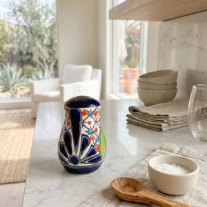 Puede incluir: Un salero de cerámica con tapa azul oscuro y diseño floral colorido. El cuerpo del salero presenta detalles en azul, naranja y verde. También se ven un pequeño cuenco de sal y una cuchara de madera.