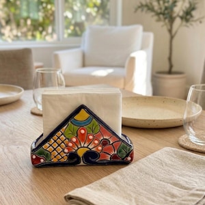 May include: A colorful, hand-painted ceramic napkin holder with a floral design in red, yellow, and green. The holder is triangular and sits on a wooden table, holding a stack of white napkins. A folded linen napkin is also on the table.
