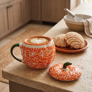 May include: A pumpkin-shaped ceramic mug with a lid, featuring a vibrant orange floral pattern on a white background and a dark green handle and stem. The mug is filled with coffee topped with heart-shaped latte art, sitting next to its lid.