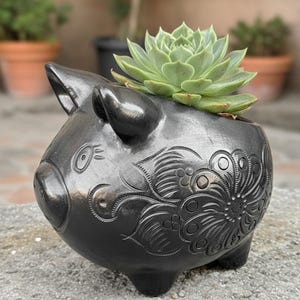 May include: Black ceramic pig planter with a succulent plant. The planter has detailed floral patterns and a glossy finish. The succulent is a light green color and adds a natural element to the decorative piece.