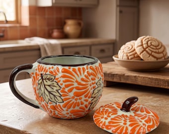 May include: A ceramic mug shaped like a pumpkin with a matching lid, featuring a vibrant orange and white floral pattern and painted green leaves. The mug has a dark brown handle and a dark brown stem on the lid, with a glossy finish throughout.