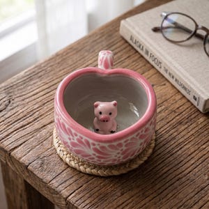 May include: A heart-shaped ceramic mug with a pink and white floral pattern on the exterior and a small, three-dimensional pink pig figurine attached to the center of the interior base. The mug features a pink handle and a smooth, glossy finish.
