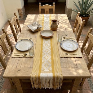 May include: A rustic wooden dining table set for a meal. The table is adorned with a yellow and white patterned runner, place settings, and a pitcher. Six wooden chairs surround the table, creating a warm and inviting atmosphere.