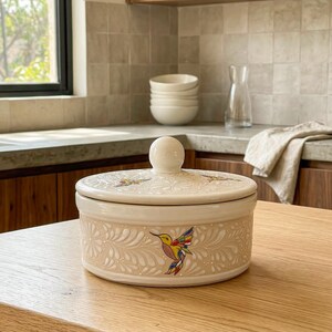 May include: A cream ceramic lidded container featuring a hummingbird design. The container has a round shape with a floral pattern and a small, round knob on the lid. The hummingbird is depicted with colourful wings.