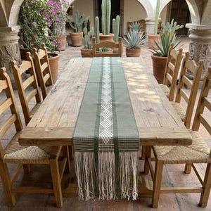 May include: A rustic wooden dining table with six matching chairs. The table is adorned with a green and white patterned table runner with fringe. The chairs have woven seats and wooden backs. The setting is outdoors.