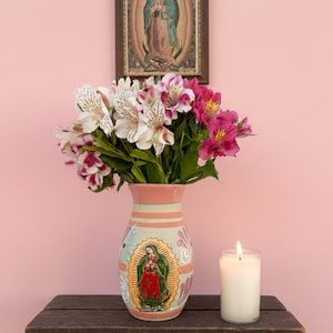 May include: A bouquet of white and pink flowers in a decorative vase with a religious image. A framed religious print hangs above the vase. A lit candle sits on a wooden shelf against a pink wall.