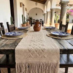 Puede incluir: Una mesa de comedor rústica está preparada para una comida. La mesa está adornada con un camino de mesa tejido beige con flecos, platos con diseños azules y blancos y una jarra marrón. Los manteles individuales tejidos y las sillas de madera oscura completan el conjunto.