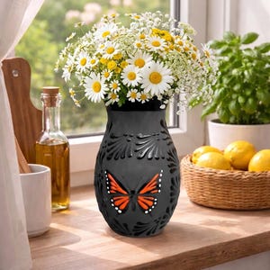 Puede incluir: Un jarrón negro con diseño de mariposa, lleno de margaritas blancas y amarillas. El jarrón está sobre una encimera de madera, con una cesta de limones y una botella de aceite al fondo.