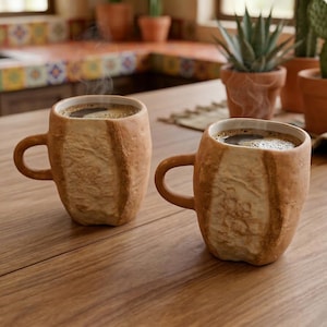 Könnte beinhalten: Zwei braune Keramikbecher mit dampfendem Kaffee auf einem Holztisch. Die Becher haben eine strukturierte, rindenartige Optik. Topfpflanzen sind im Hintergrund zu sehen, was auf eine gemütliche Küchenumgebung hindeutet.