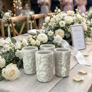 Puede incluir: Decoración de mesa para una boda con cuatro tazas de cerámica verde claro con un diseño floral blanco. La mesa está adornada con rosas blancas, vegetación y una tarjeta de menú. La escena sugiere una celebración.