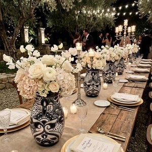 May include: A long wooden dining table set for an outdoor event, featuring black and white patterned ceramic vases filled with white and cream floral arrangements, silver candelabras, gold-rimmed plates, and white napkins with printed text.