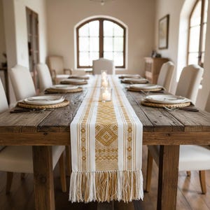 Op de afbeelding: Een rustieke houten eettafel met een wit en goudkleurig patroon tafelloper met franjes. De tafel is gedekt met borden, placemats en kaarsen, wat een warme en uitnodigende sfeer creëert.