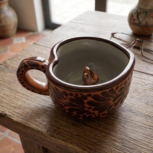 May include: A heart-shaped ceramic mug with a brown and terracotta floral pattern. The exterior features a dark brown handle with a spotted design. Inside the mug, a small, sculpted brown animal figure sits at the bottom, visible against the light interior.