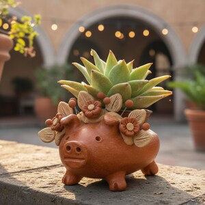 May include: A terracotta pig-shaped planter with a succulent plant. The planter has a detailed design with floral accents and is filled with a green succulent. The background includes other potted plants and a blurred outdoor setting.
