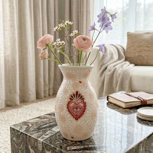 May include: A white ceramic vase with a heart and eye design, filled with pink and purple flowers. The vase is on a marble table, with a neutral-toned background.