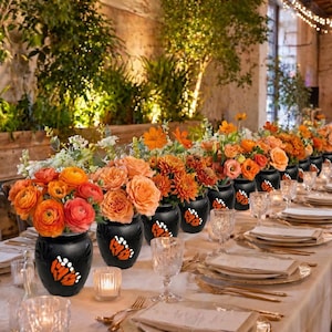 May include: A long table set for a formal event, featuring black vases with butterfly designs filled with orange and peach flowers. The table is set with gold-rimmed plates, crystal glasses, and candles, creating an elegant ambiance.