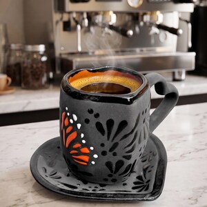 May include: A black ceramic coffee cup and saucer set with a butterfly design in orange and white. The cup is filled with hot coffee, and steam rises from the top. The cup and saucer are on a marble countertop.