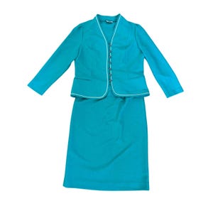 May include: A teal two-piece skirt suit. The jacket has long sleeves, a V-neck, and a row of buttons down the front. White trim outlines the jacket and skirt. The skirt is a straight, knee-length style.