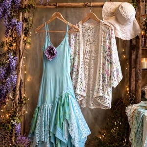May include: A light blue spaghetti strap dress with a purple flower accent and a layered, asymmetrical hem featuring lace and fabric panels, paired with a white floral lace cardigan and a white sun hat hanging on wooden hangers.