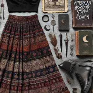 May include: A black lace-trimmed crop top and a long, patterned skirt with red, brown, and blue details are displayed. Accessories include necklaces, rings, a dagger, and books with the title "American Horror Story Coven". Black ankle boots complete the ensemble.