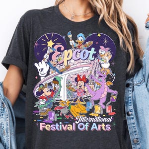 May include: Dark gray t-shirt featuring a colorful graphic of Disney characters, including Mickey Mouse, Minnie Mouse, and Figment, with the text "Epcot International Festival of Arts". The design includes various art supplies.