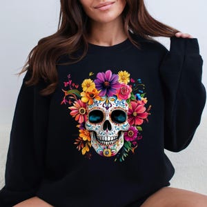 Op de afbeelding: Zwarte sweatshirt met een kleurrijk suikerschedelontwerp. De schedel is versierd met ingewikkelde bloempatronen en omgeven door een boeket van heldere bloemen in roze, geel, paars en oranje. Het ontwerp is gecentreerd op de voorkant.
