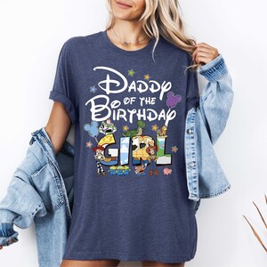 A woman wearing a blue t-shirt with a Disney-themed design and text, standing in front of a white wall.